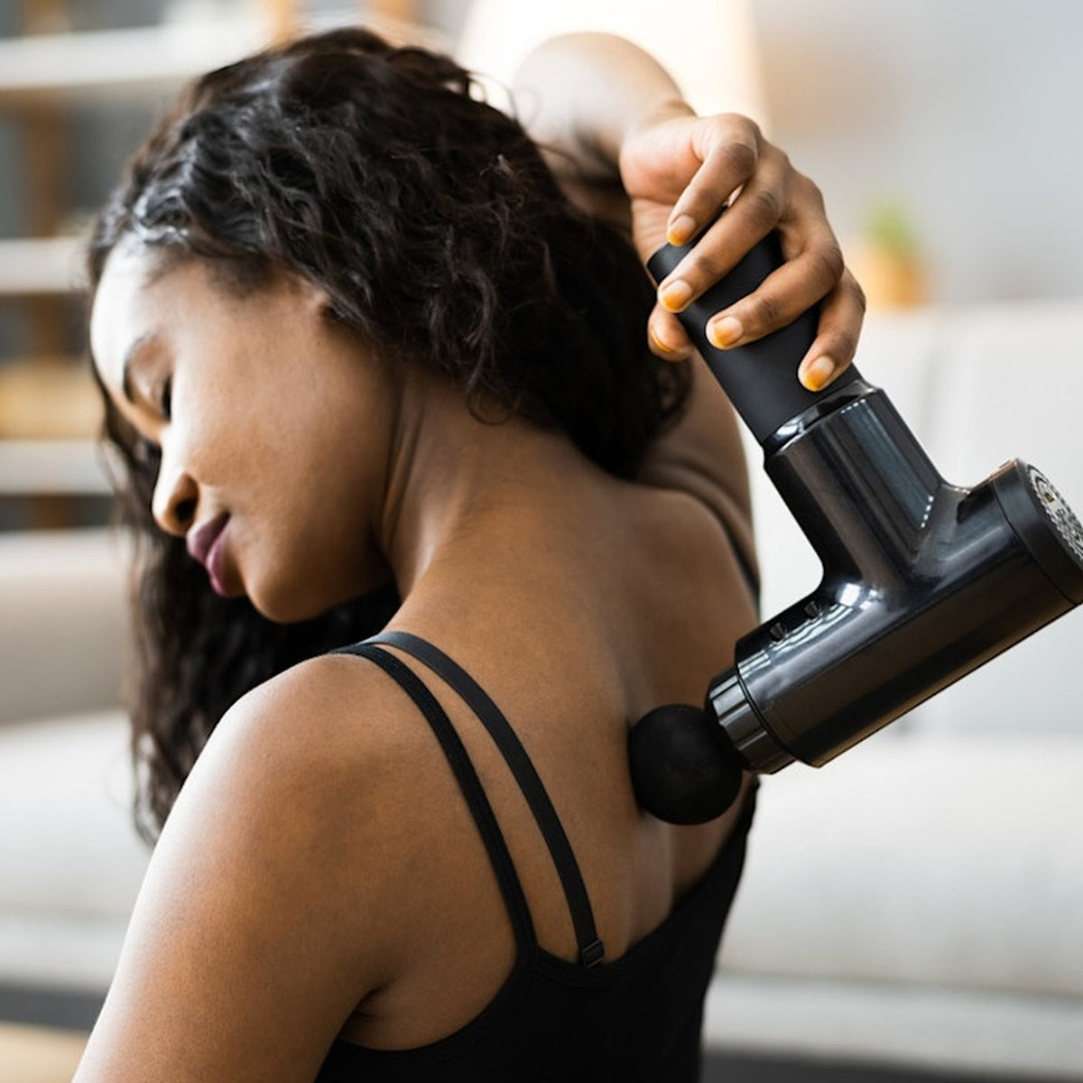 woman using massage gun on shoulder for muscle pain relief at home